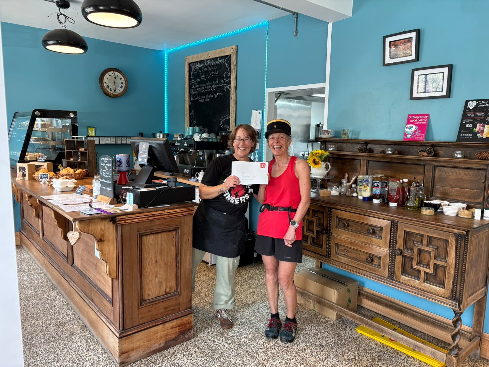 Two smiling women hold up a delivered letter at the café’s old wooden post office counter, with the old post office clock on the wall behind.