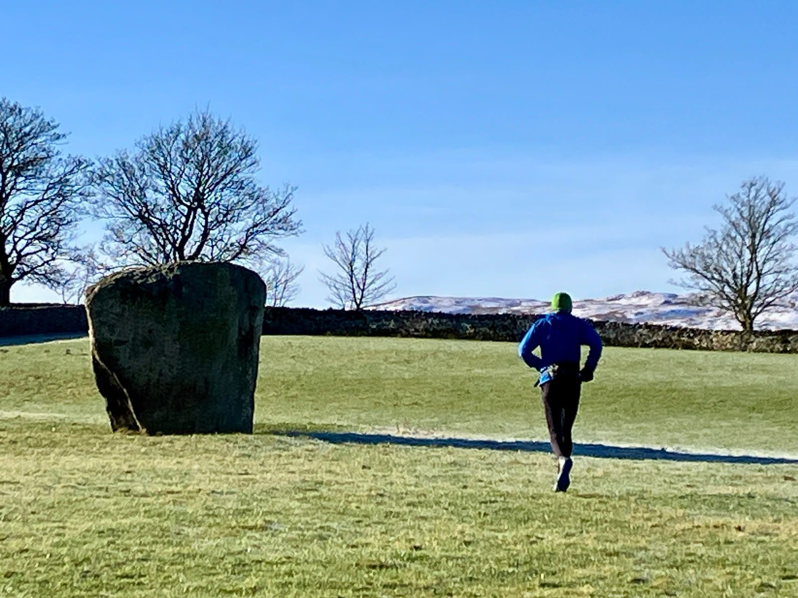 A runner passes a huge standing stone in a frosty field under a blue sky.