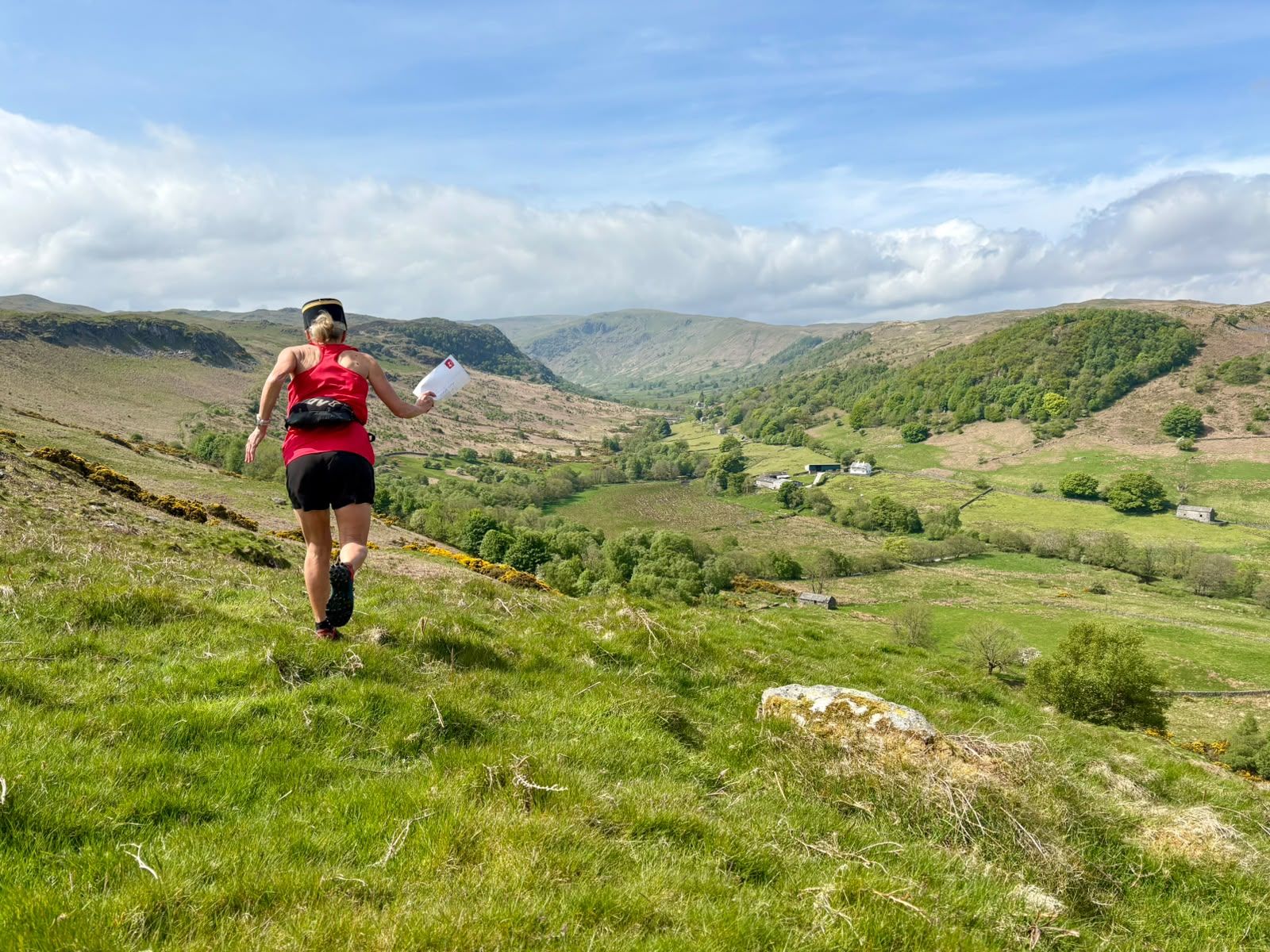 A runner in a red vest and postie’s cap bounds down a green fellside holding a letter, with a wide valley of farms and woods below.