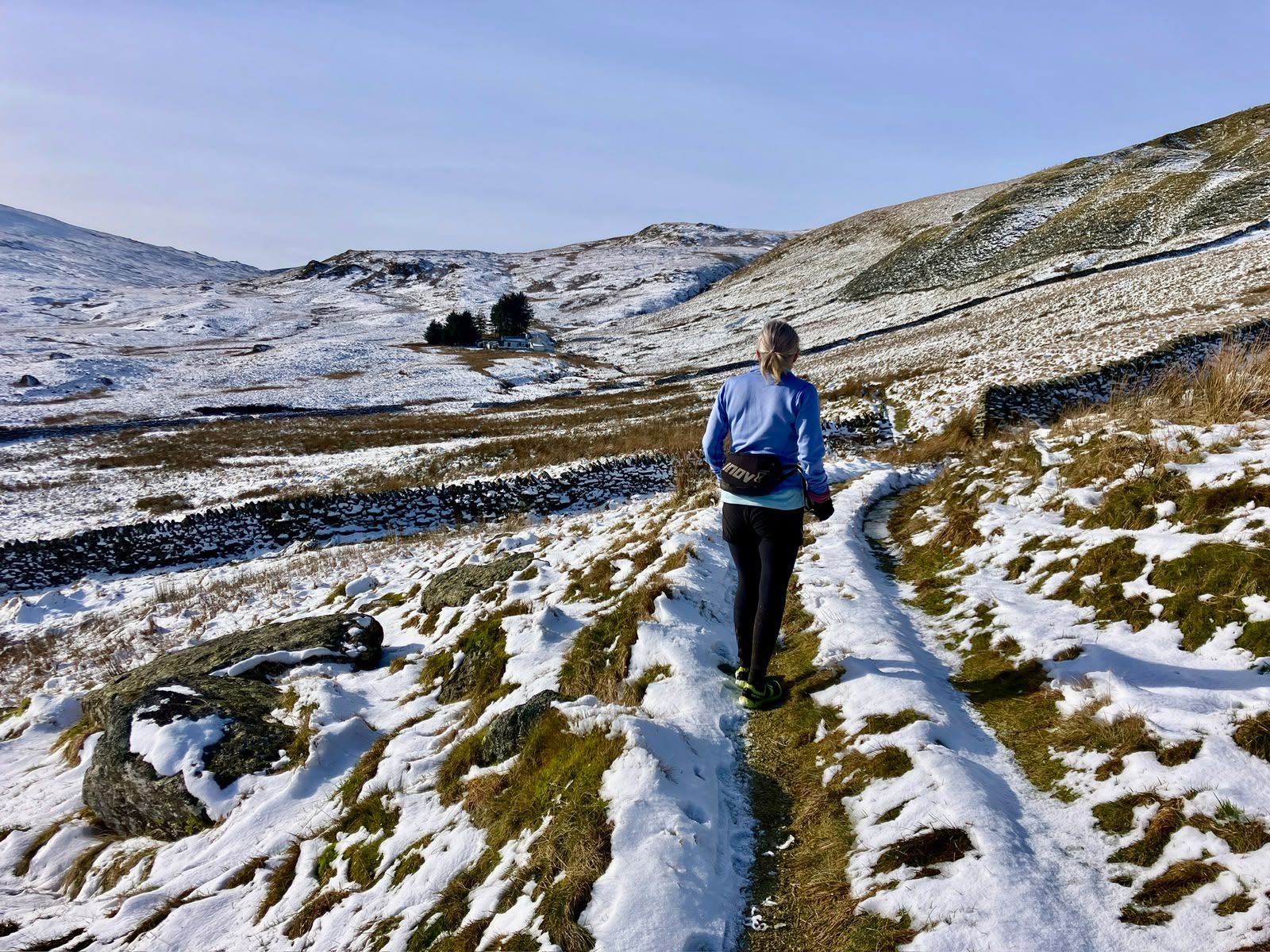 A walker follows a snowy track between stone walls into a white valley.